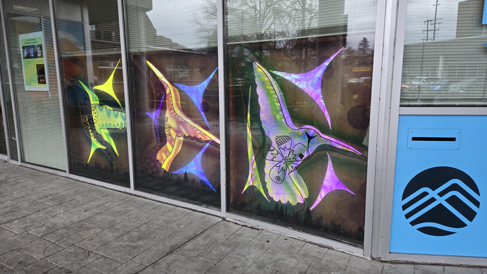 Three giant artistic hummingbirds made by artist Rain Pierre glow through a window, shining a variety of vibrant neon colours, orange, blue, green, yellow, pink and purple.